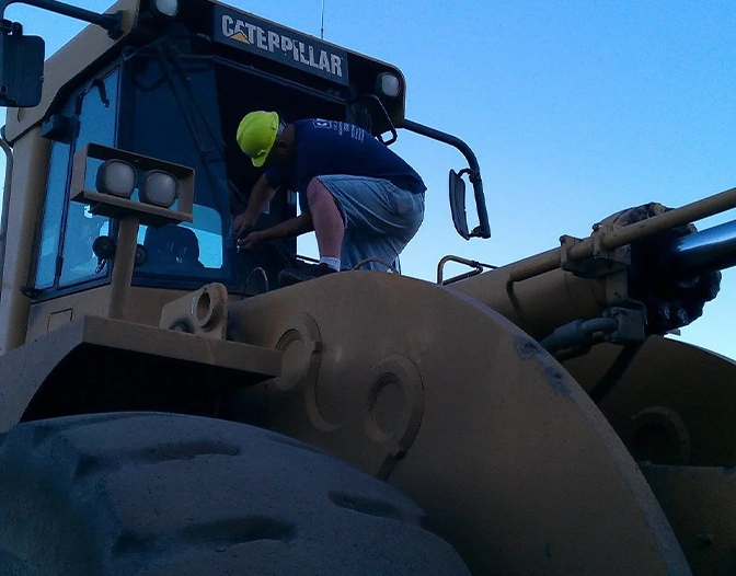 A person in a yellow helmet stands on a large Caterpillar construction vehicle, handling a Rig Window Replacement near the front window at dusk.