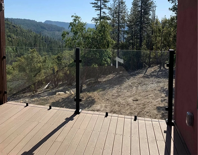 A wooden deck with a sleek deck glass railing overlooks a dry, wooded landscape with trees and hills in the background.