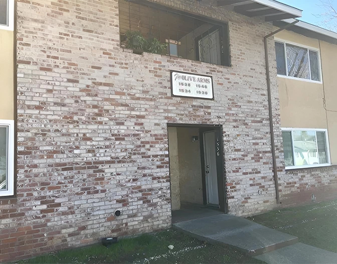 A two-story brick apartment building with an open doorway and a sign above reading "Olive Arms Apartments" displays its street numbers.