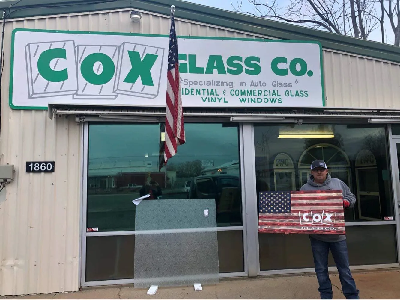 A person stands outside Cox Glass Co., holding a flag-themed sign. A glass pane and an American flag are displayed in front of the building.