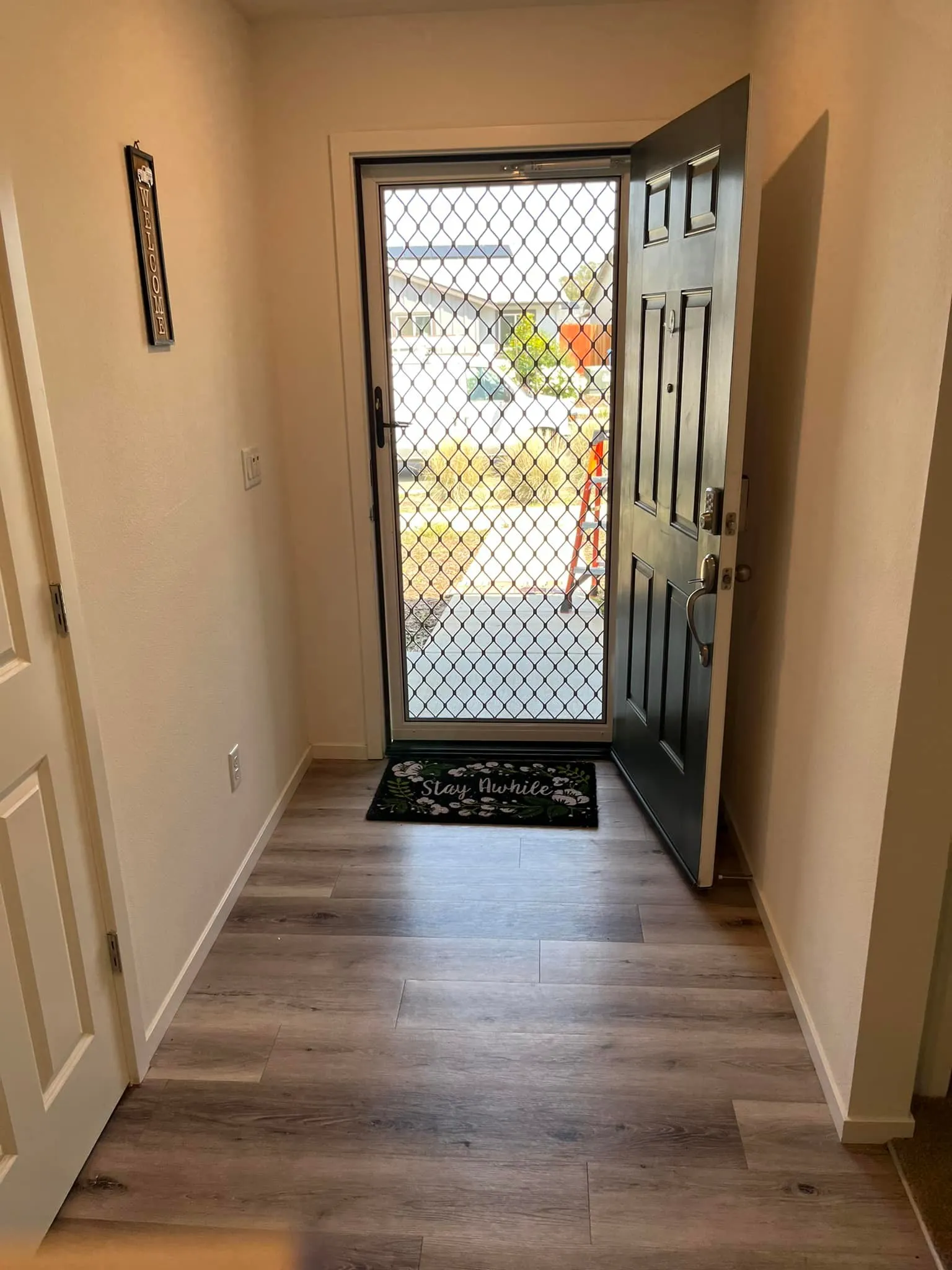 A hallway with light wood flooring, an open black front door with a mesh screen, and a doormat that reads "Stay Humble." Sunlight and outdoor scenery are visible through the door.
