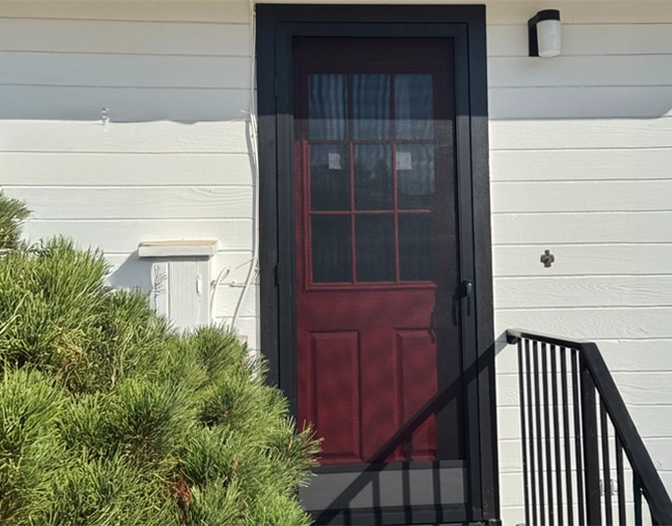 A red and black door with a screen is set in a white exterior wall; a railing and green shrub are visible in the foreground.