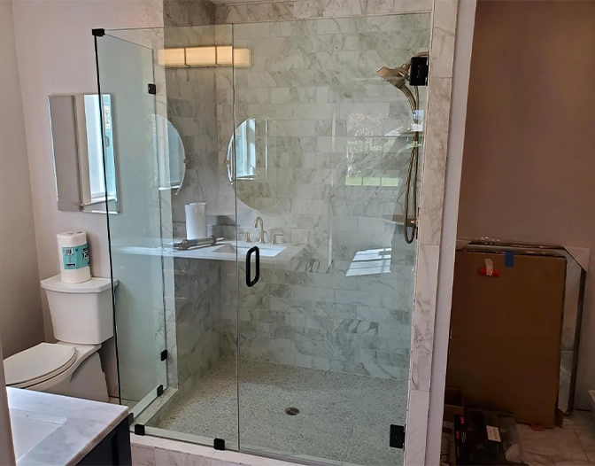 Glass-enclosed, modern shower enclosure with marble tile walls, black hardware, and built-in showerhead; adjacent countertop, toilet, and window are visible.