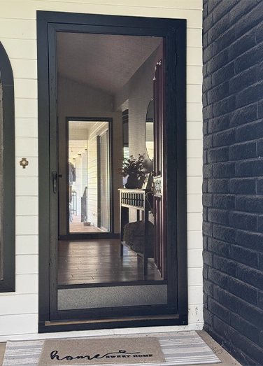 A black-framed glass storm door reveals a hallway with a console table, flowers, and mirrors; a "home sweet home" doormat lies in front.