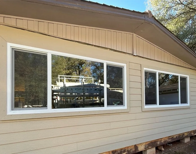 A tan house exterior features three large residential windows with white frames, reflecting trees and a vehicle; the house is elevated on blocks with unfinished ground below.
