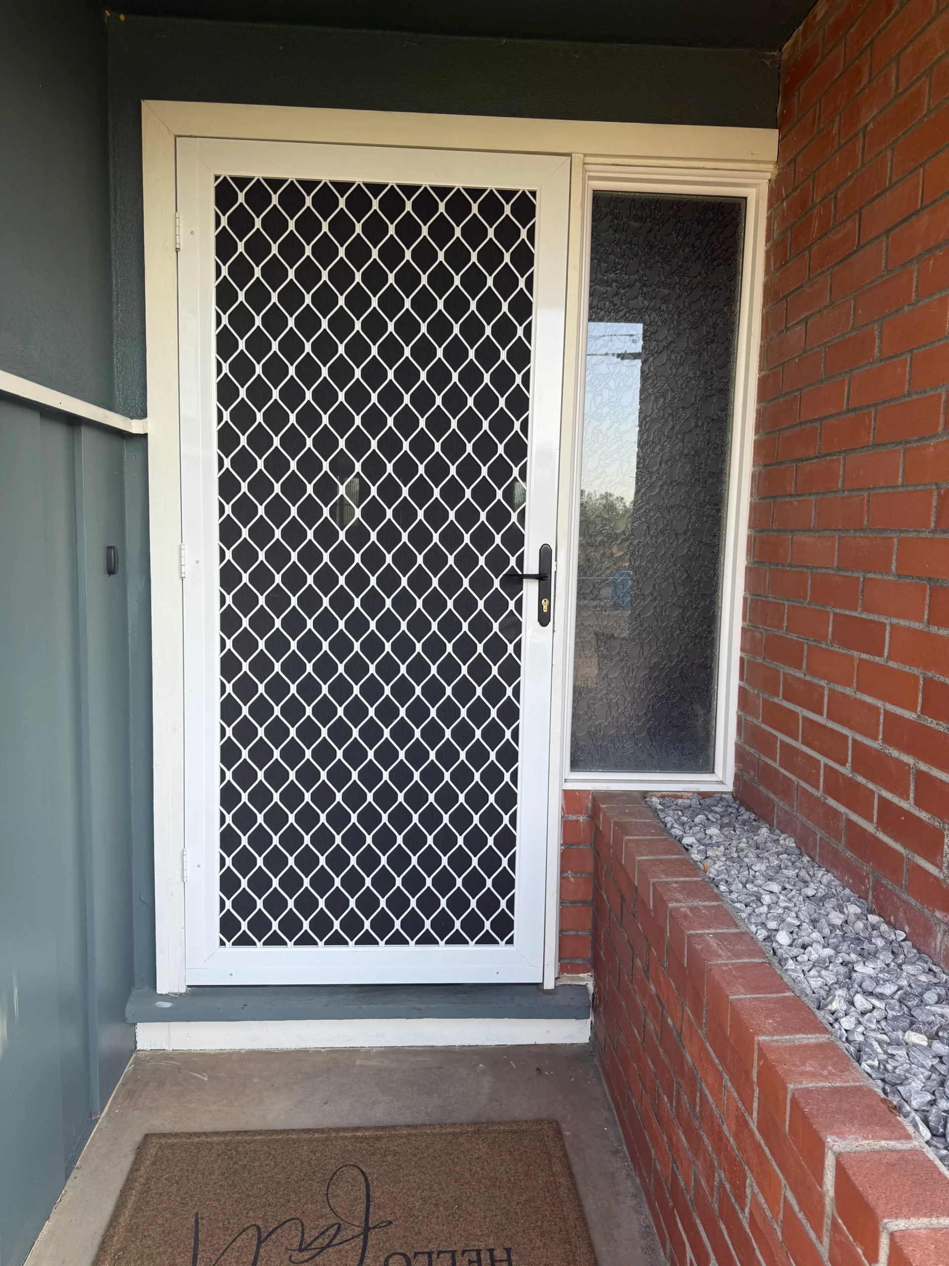 A white security screen door with a diamond mesh pattern and a glass panel beside it. A “hello” doormat lies on the concrete entrance, next to a brick planter with gray rocks.