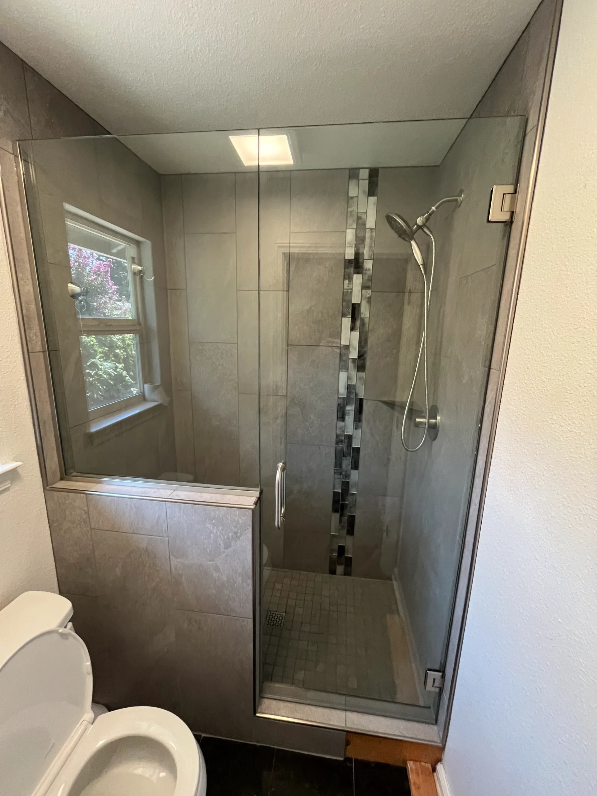 Modern walk-in shower with glass door, wall-mounted showerhead, vertical accent tile strip, and adjacent window; toilet is partially visible in the foreground.