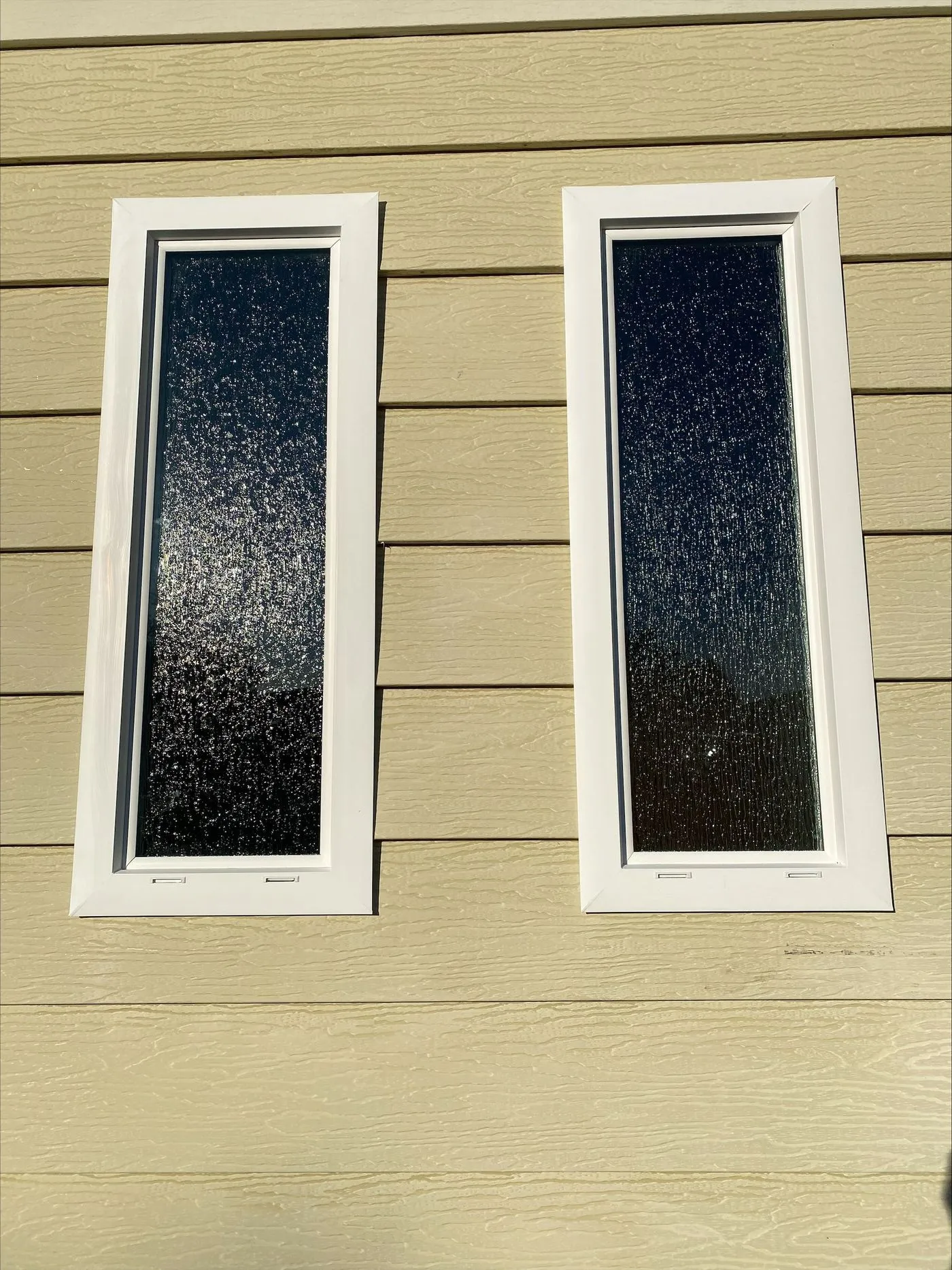 Two narrow rectangular windows with white frames set into a beige horizontal siding wall, shown in sunlight.