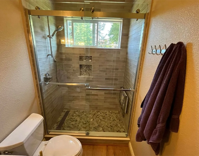 A tiled walk-in shower with a frameless shower door, wall-mounted showerhead, built-in shelves, and a window above. A towel hangs on a hook, and a toilet is visible in the foreground.