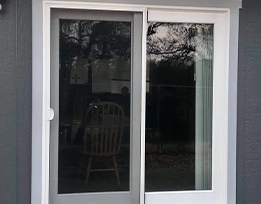 A white-framed sliding glass door with a wooden chair visible inside and outdoor trees reflected in the glass.
