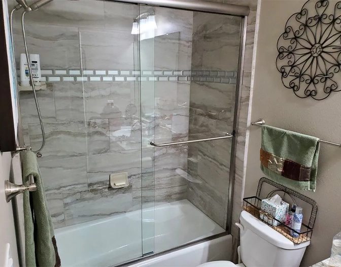 A bathroom with a glass-enclosed bathtub and shower, tiled walls, a towel rack, a toilet with a storage basket, and a decorative metal wall piece.