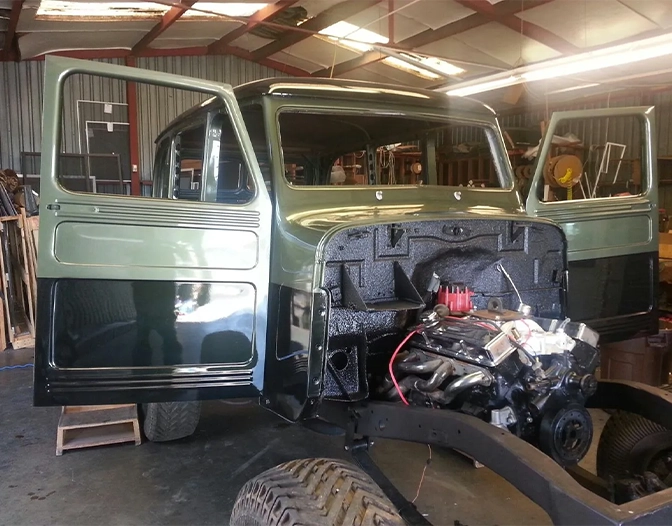 A partially restored vintage pickup truck sits in a workshop, with its hood removed and engine exposed, and both front doors open.