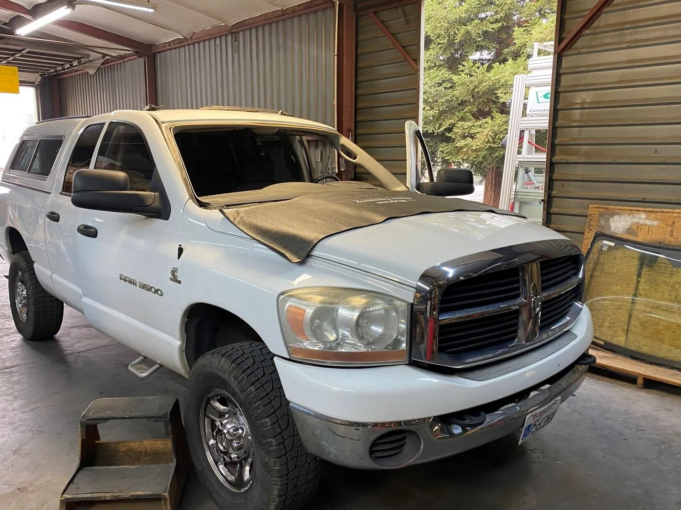 A white Dodge Ram 2500 pickup truck with a damaged windshield and hood sits inside a garage, with the driver's door open and a step platform beside it.