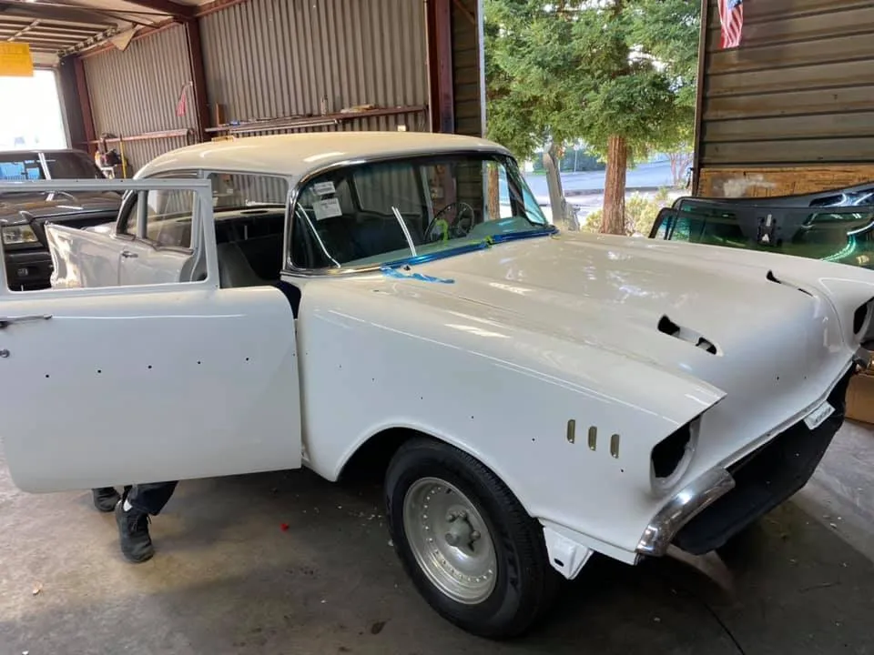 A white classic car with its driver door open is parked inside a garage; a person is partially visible behind the door.