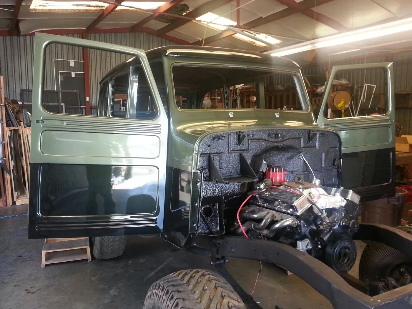 A partially restored vintage pickup truck with its hood and front body panels removed, exposing the engine, parked inside a workshop with doors open.