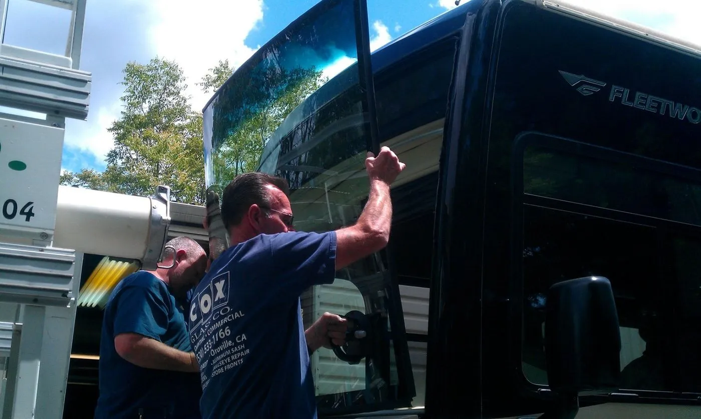 Two workers are installing or replacing a large windshield on a black Fleetwood RV using suction cup tools.