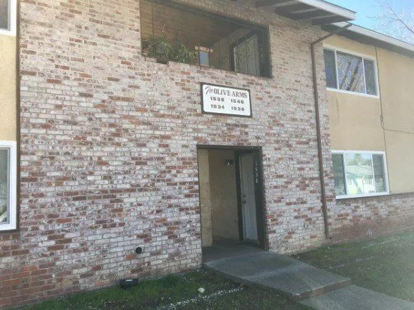 Front entrance of a two-story brick apartment building with a sign reading "The Olive Arms" and apartment numbers above an open doorway.