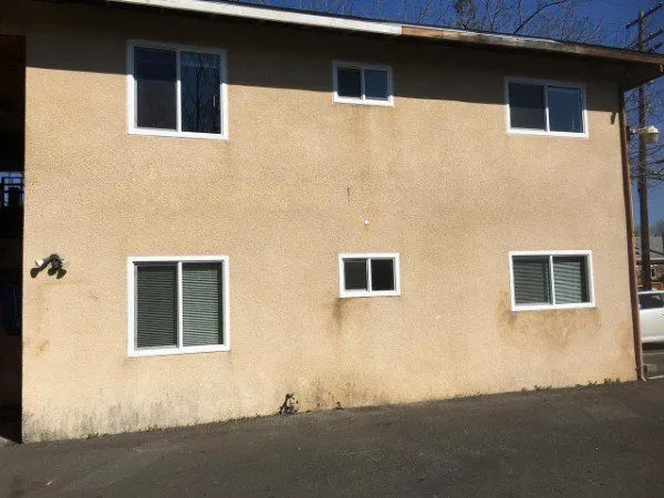 A beige building wall with five white-framed windows of varying sizes, and a water faucet on the lower left side, facing an empty paved area.