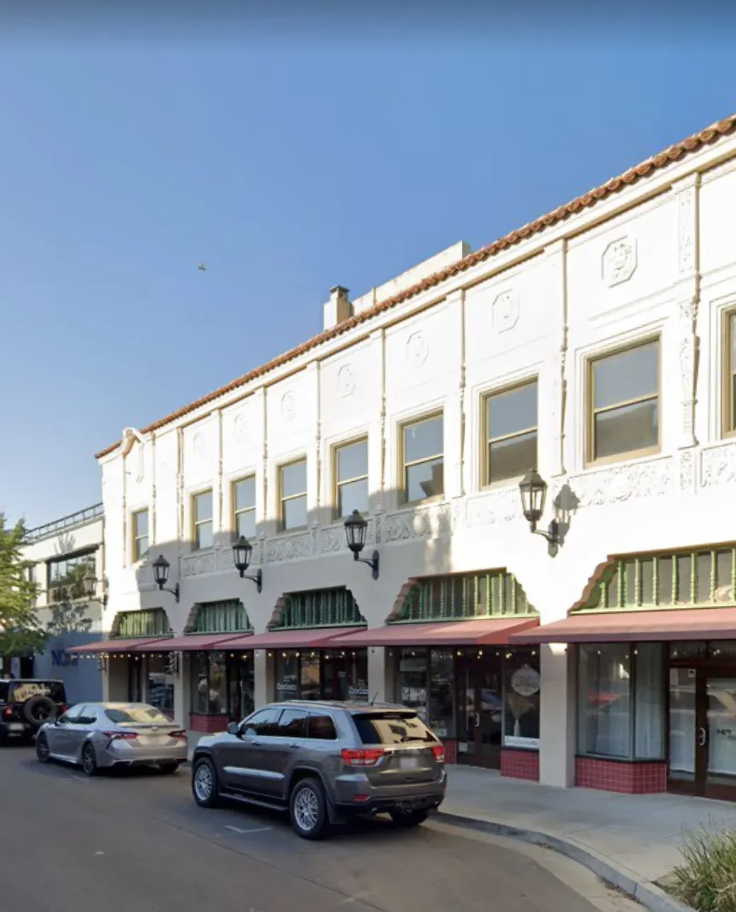 A street view shows parked cars in front of a two-story white building with balconies and storefronts on the ground floor.