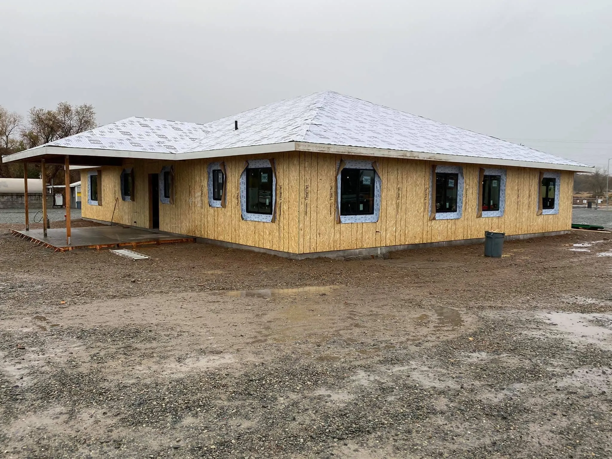 A single-story building under construction with unfinished wooden walls, covered windows, a covered porch, and a shingled roof on a muddy, gravel lot.