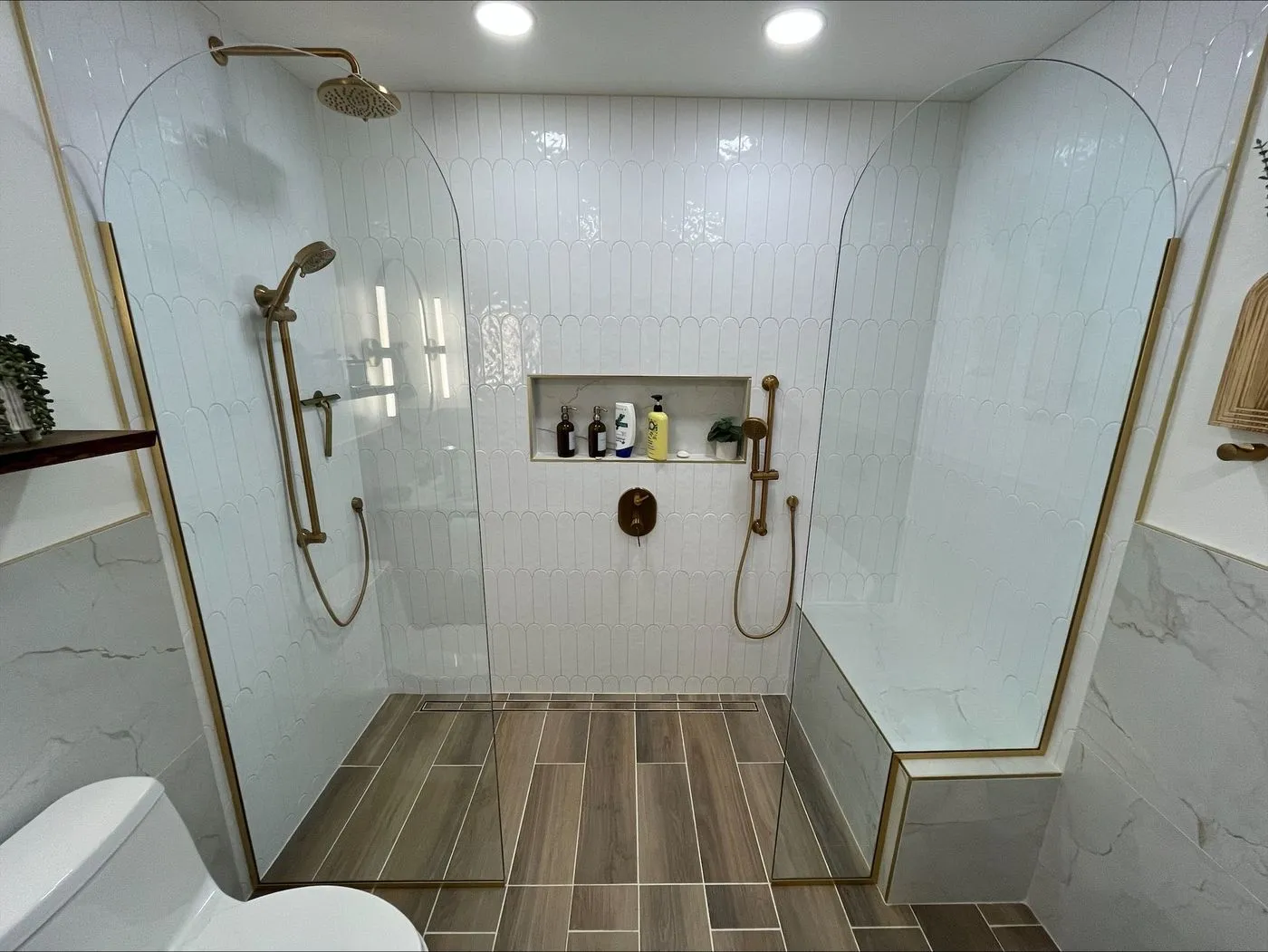 Modern double shower with white tile walls, gold fixtures, built-in bench, recessed shelf with toiletries, and wood-look tile flooring.