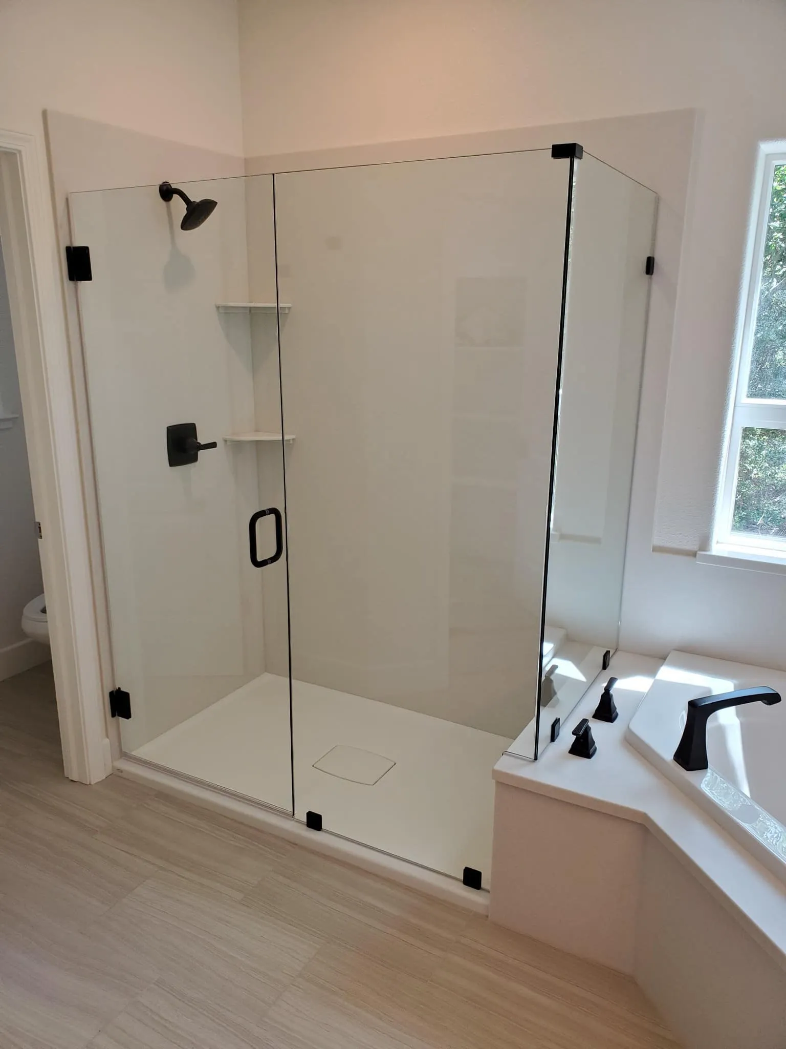 A modern bathroom with a glass-enclosed corner shower, built-in shelves, black fixtures, and a white bathtub next to a window.