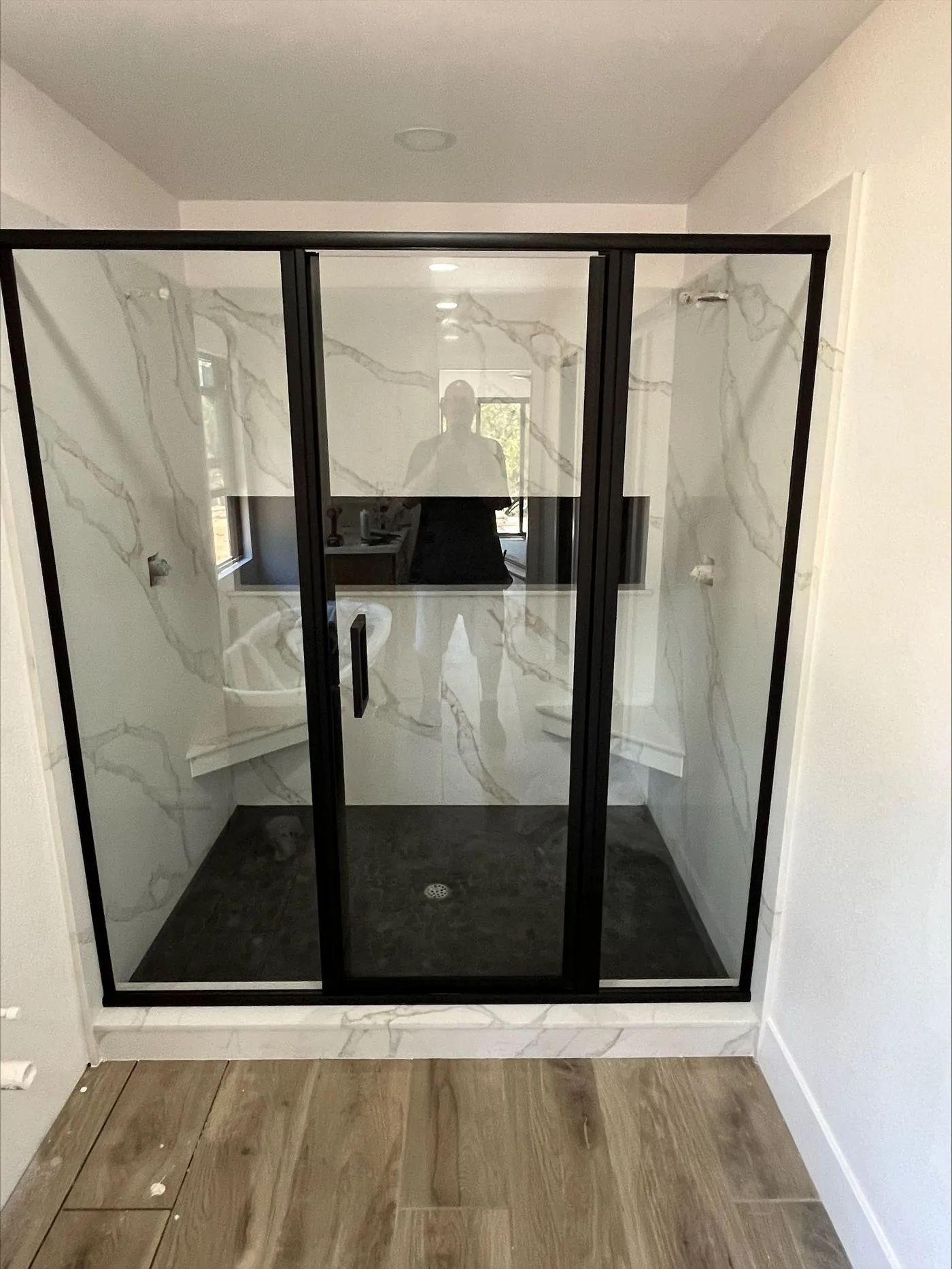 A modern walk-in shower with black-framed glass doors, marble-patterned walls, built-in bench seating, and light wood flooring outside.