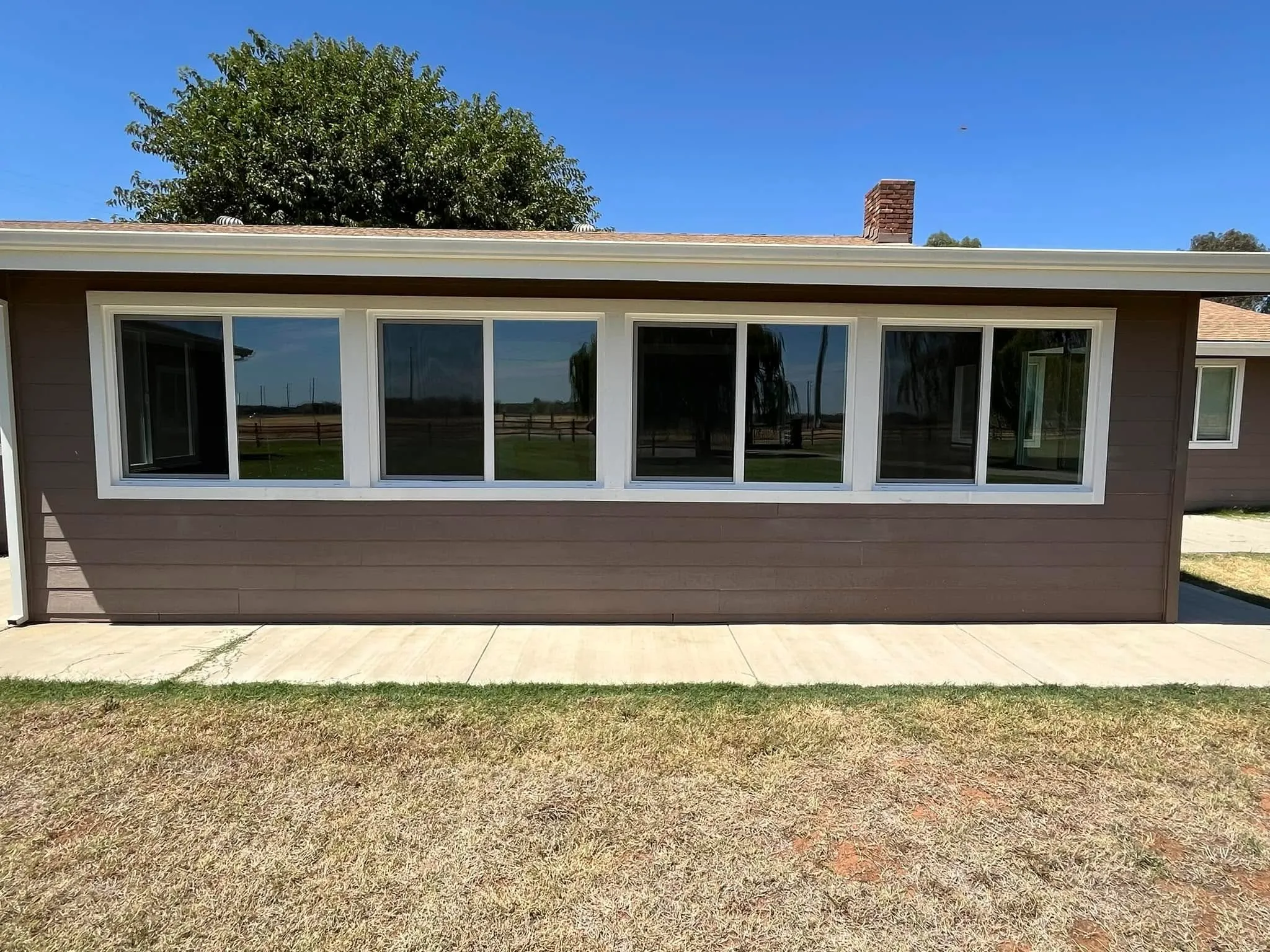 A single-story house with brown siding and large rectangular windows facing a concrete walkway and dry grass lawn under a clear blue sky.
