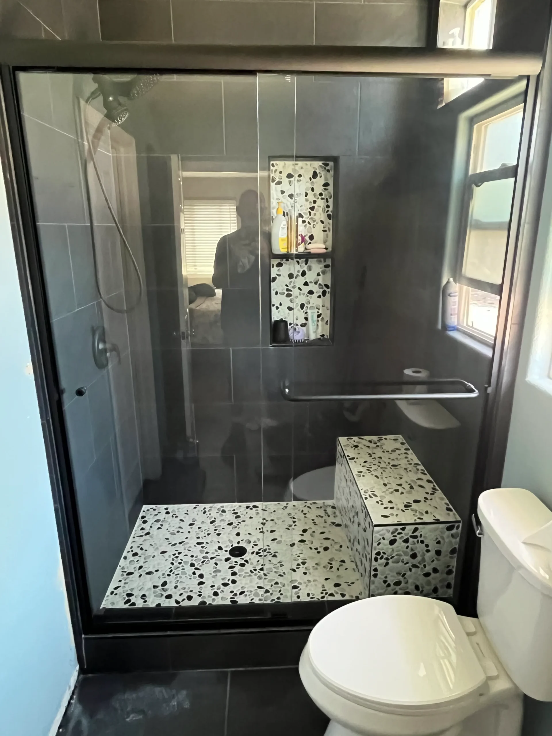 Modern bathroom with a glass-enclosed shower featuring terrazzo tile bench and shelves, a wall-mounted showerhead, and a white toilet in the foreground.