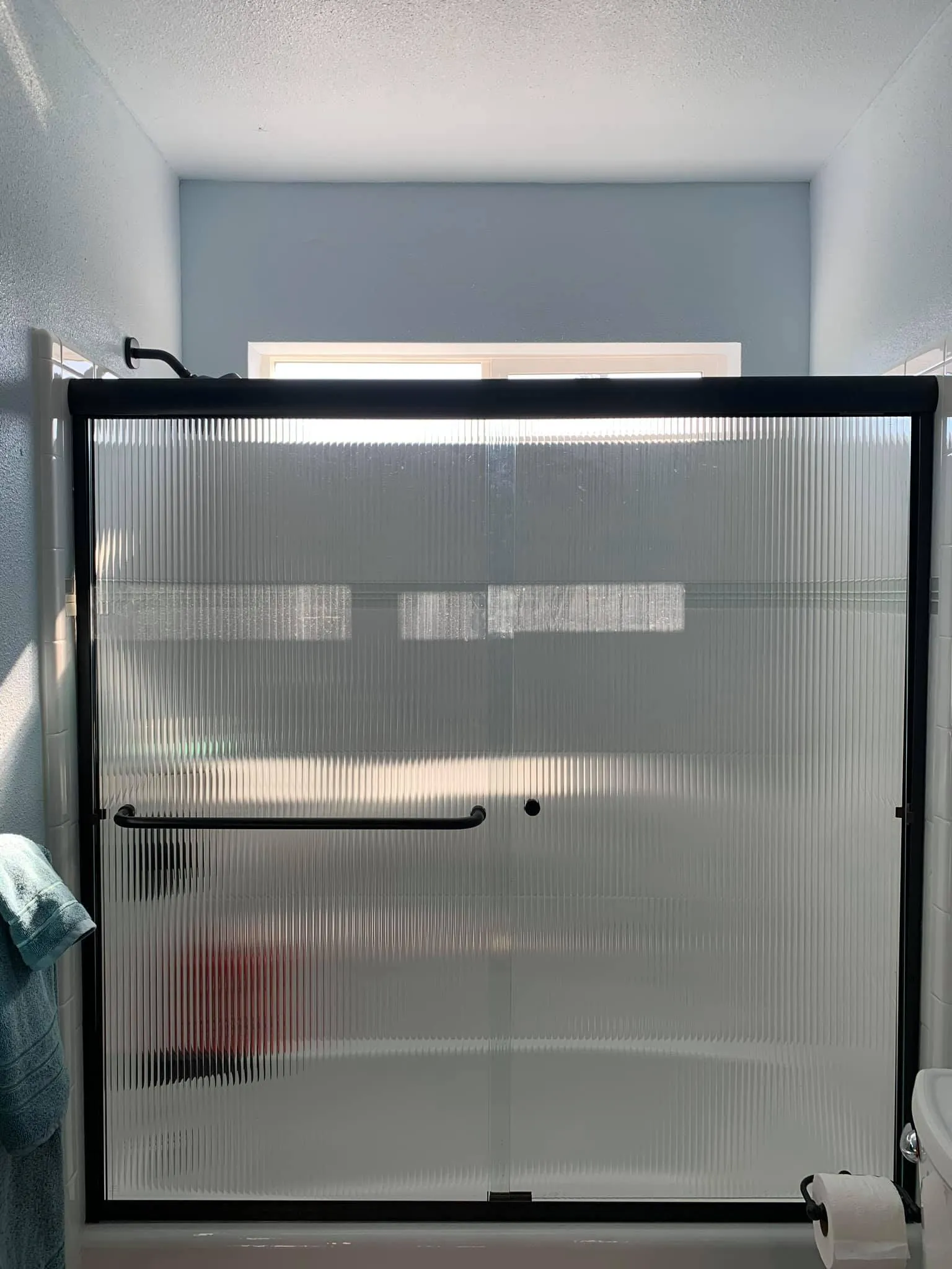 A shower with frosted sliding glass doors, black metal frame, and towel hanging on a nearby rack, illuminated by sunlight from a window above.