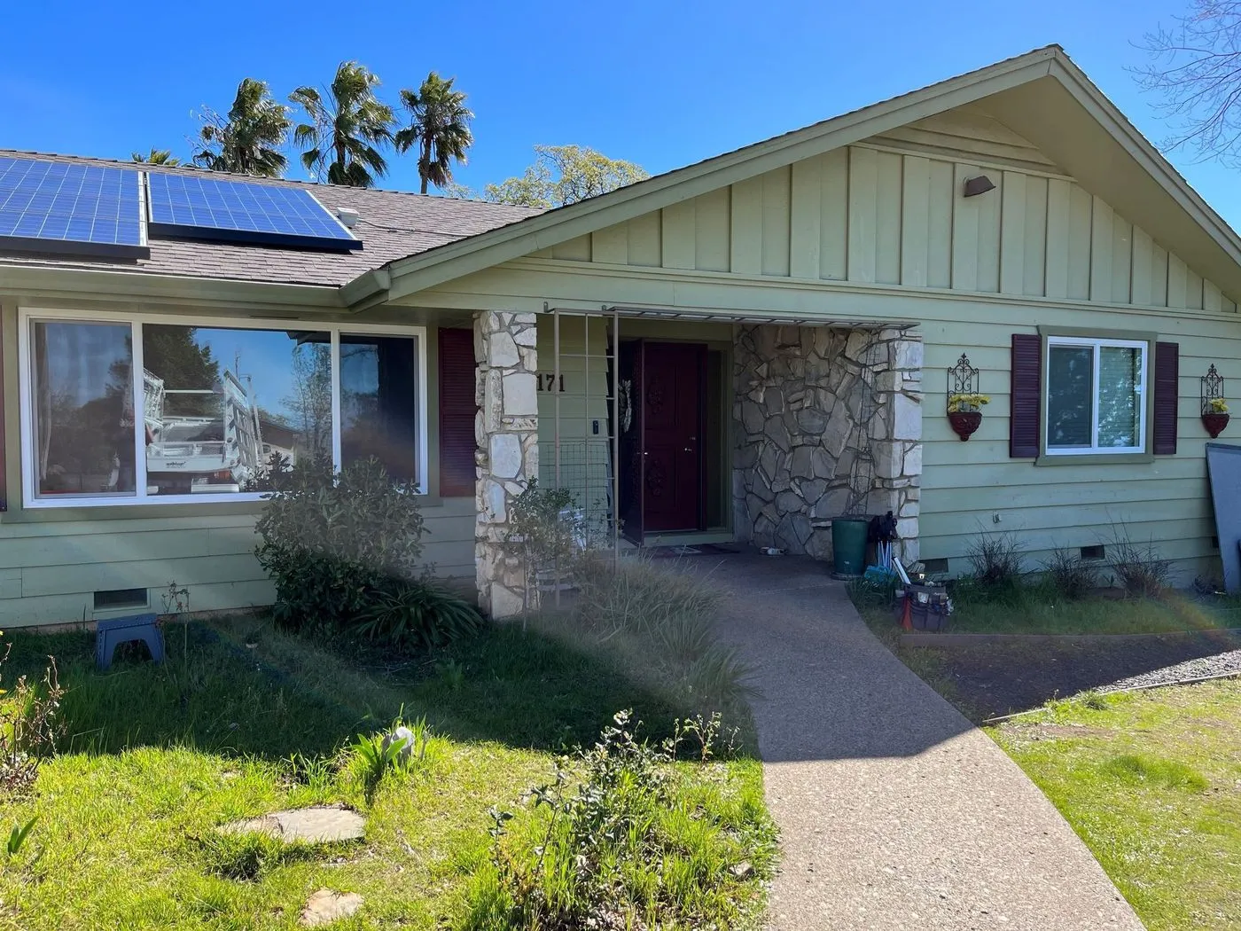 Single-story house with light green siding, stone accents, solar panels on the roof, and a covered front entrance, set on a sunny day.