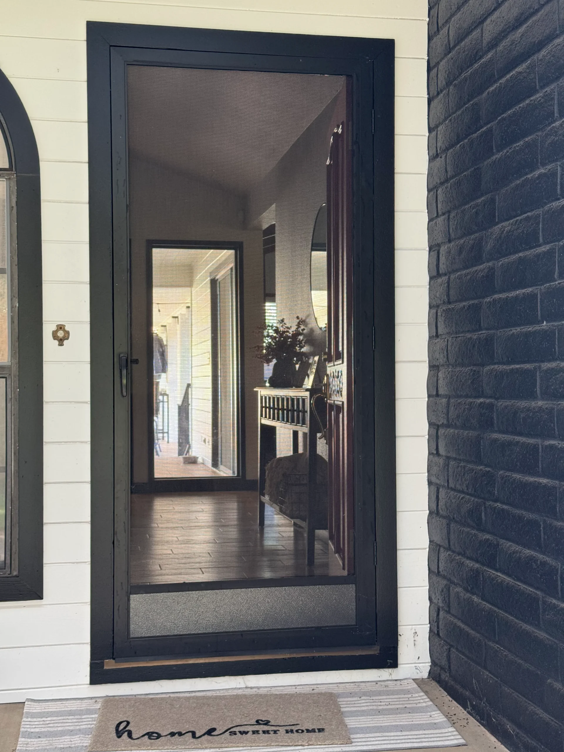 A glass storm door with a black frame reflects a porch and entryway interior. A doormat reading "home sweet home" is placed on a striped rug in front of the door.