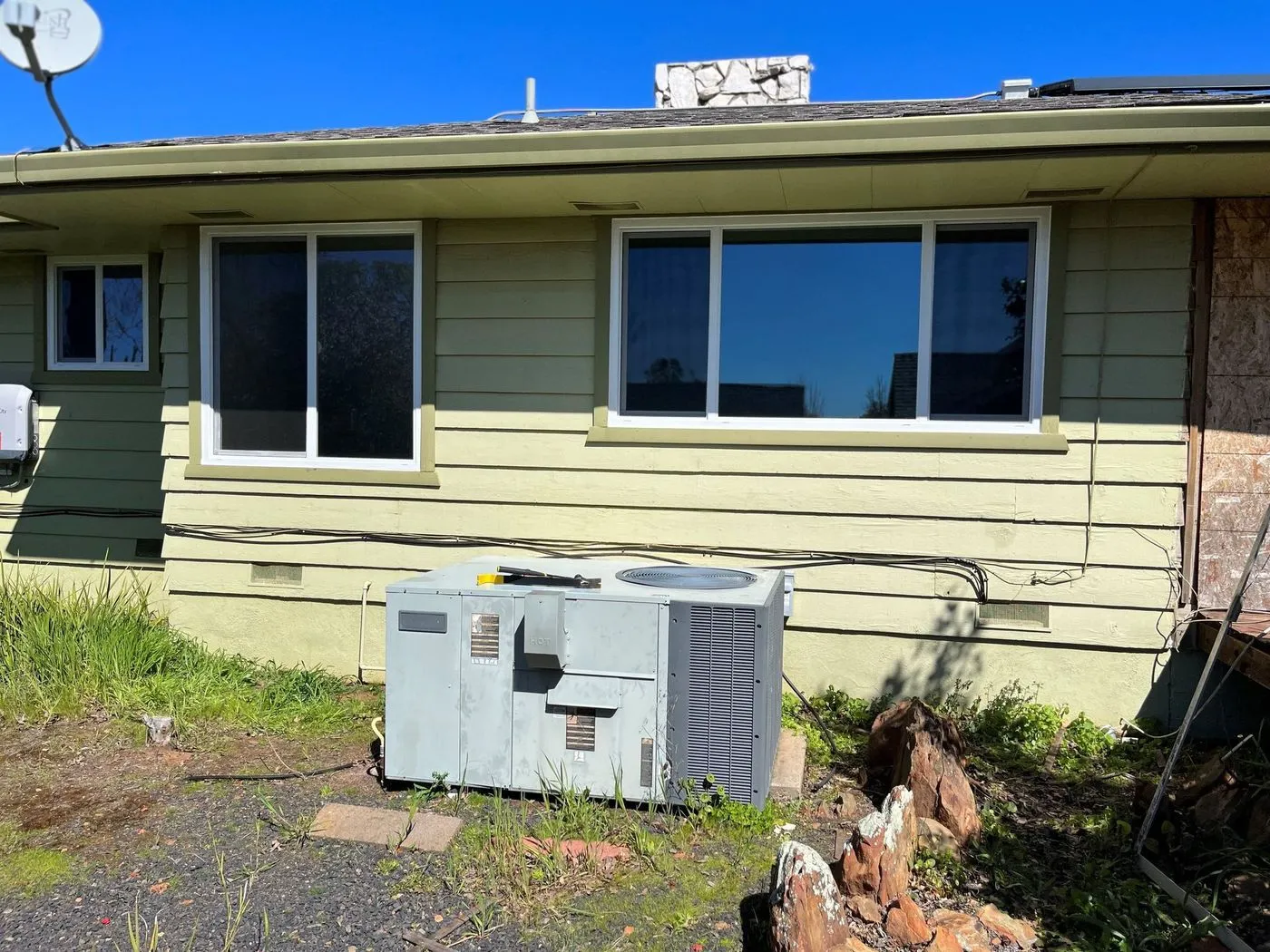 A central air conditioning unit is installed on the ground outside a green single-story house with two large windows.
