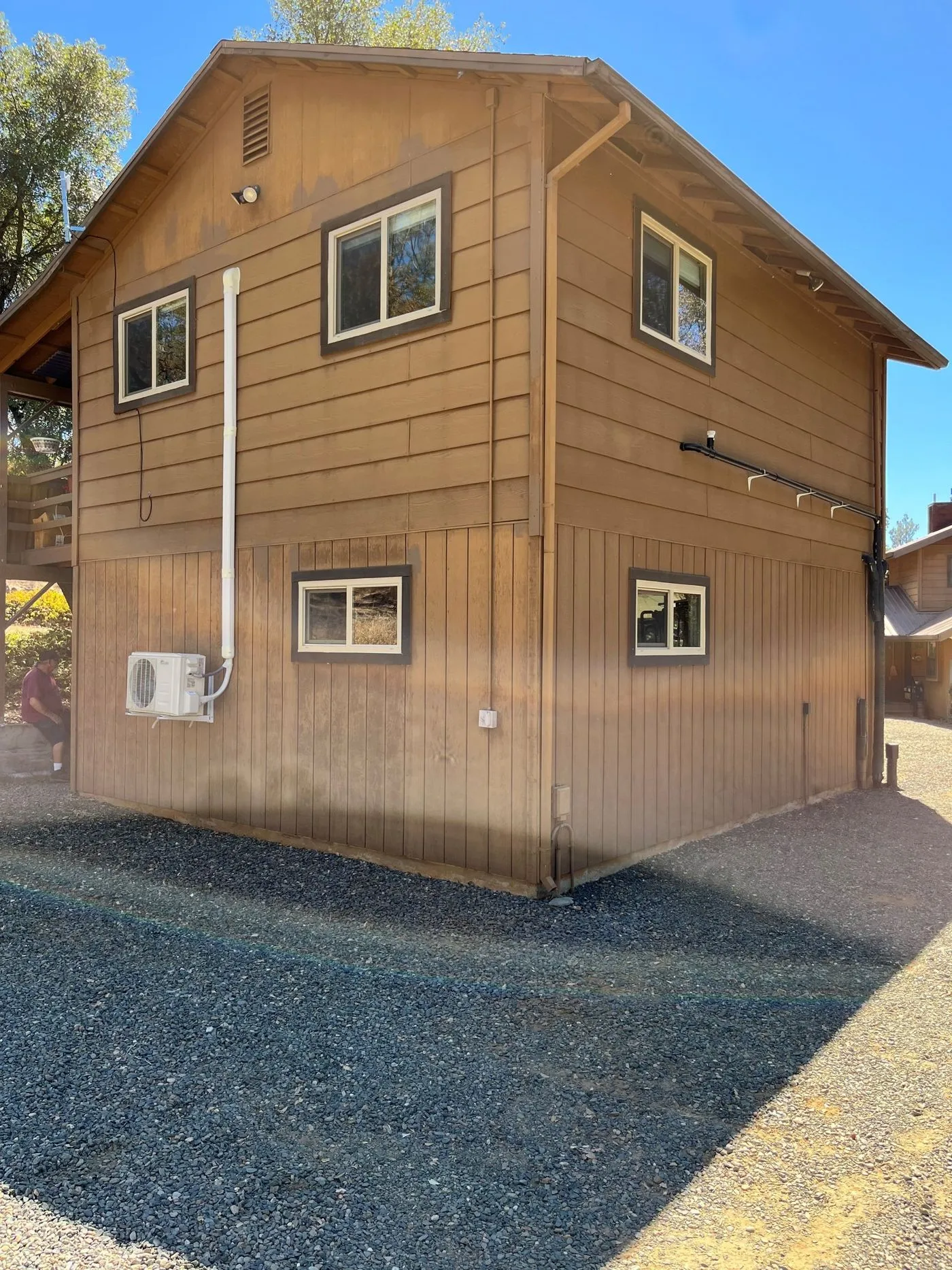 A two-story brown wooden building with several windows, an exterior air conditioning unit, and gravel surrounding the structure on a sunny day.