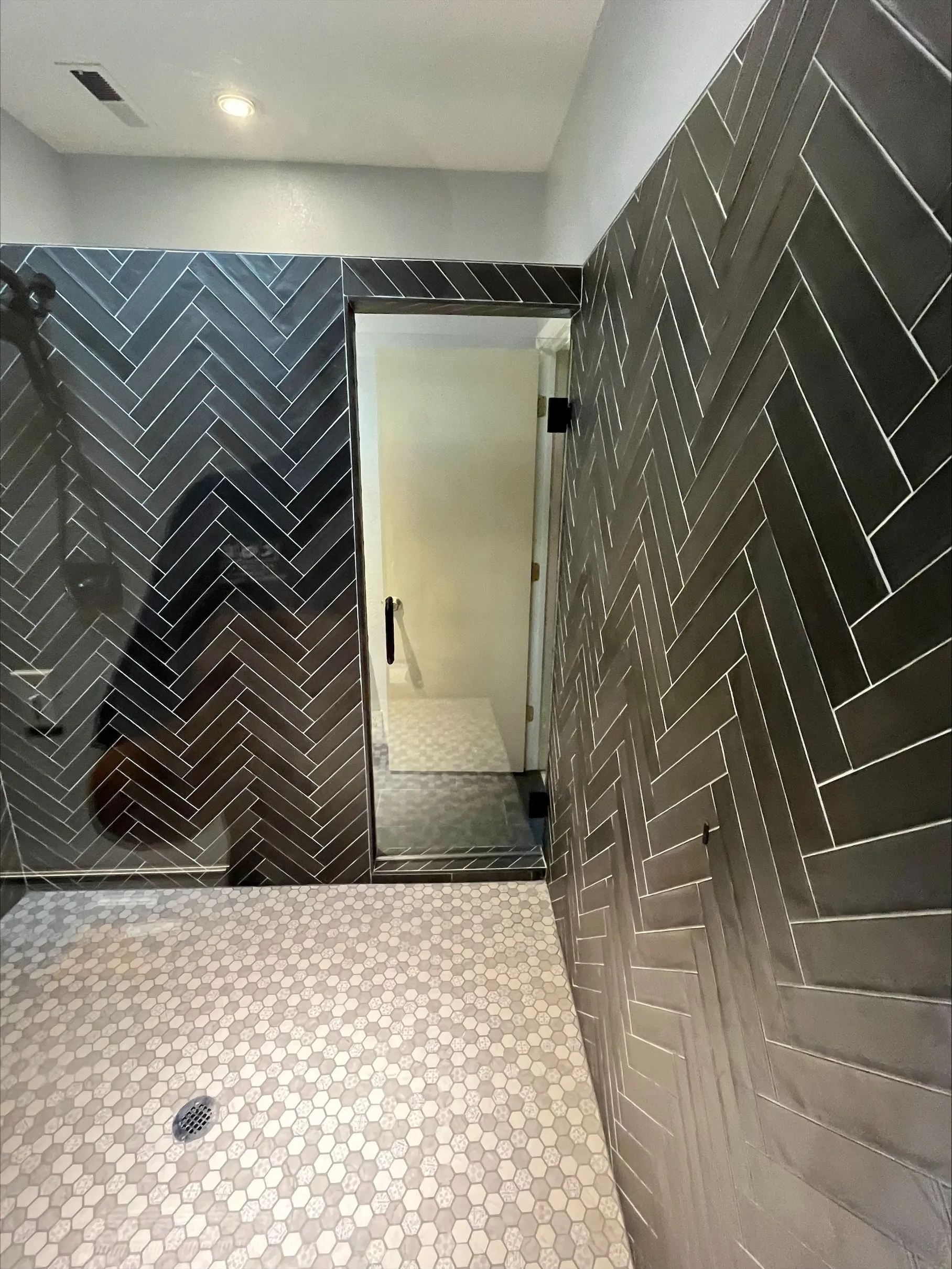 A bathroom with dark herringbone-patterned tiled walls, a hexagonal tile floor, a glass shower door, and a recessed ceiling light.