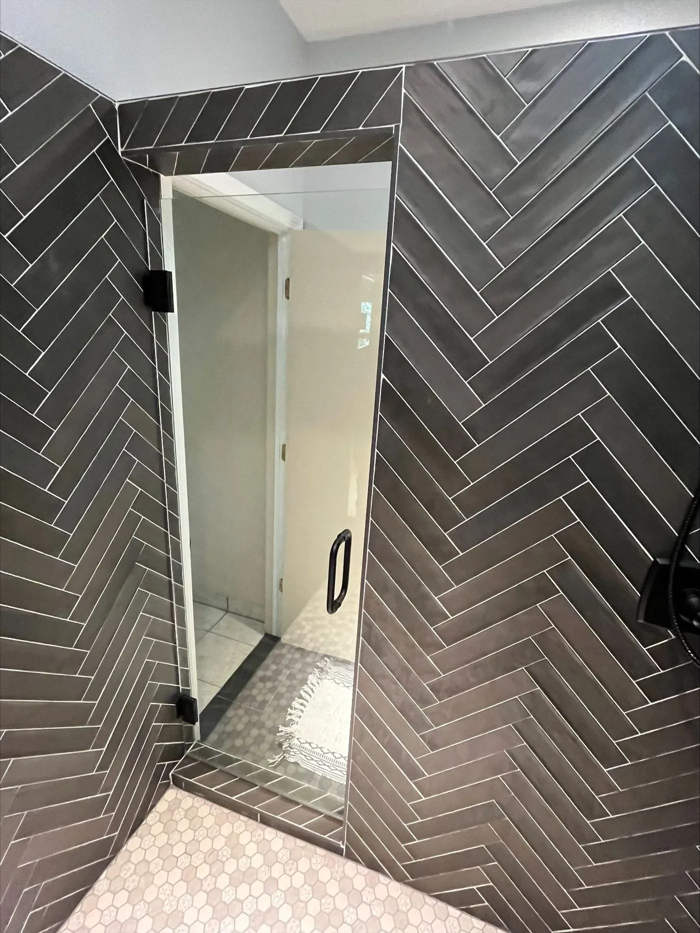 Glass shower door with black handle set in a bathroom with dark herringbone tile walls and hexagonal floor tiles.