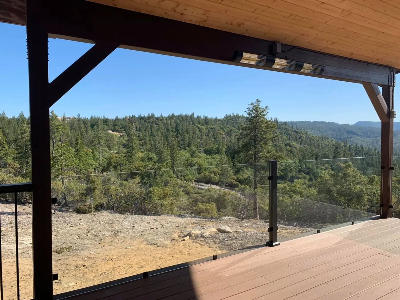 View from a covered wooden deck with glass railing, overlooking a forested landscape and hills under a clear blue sky.