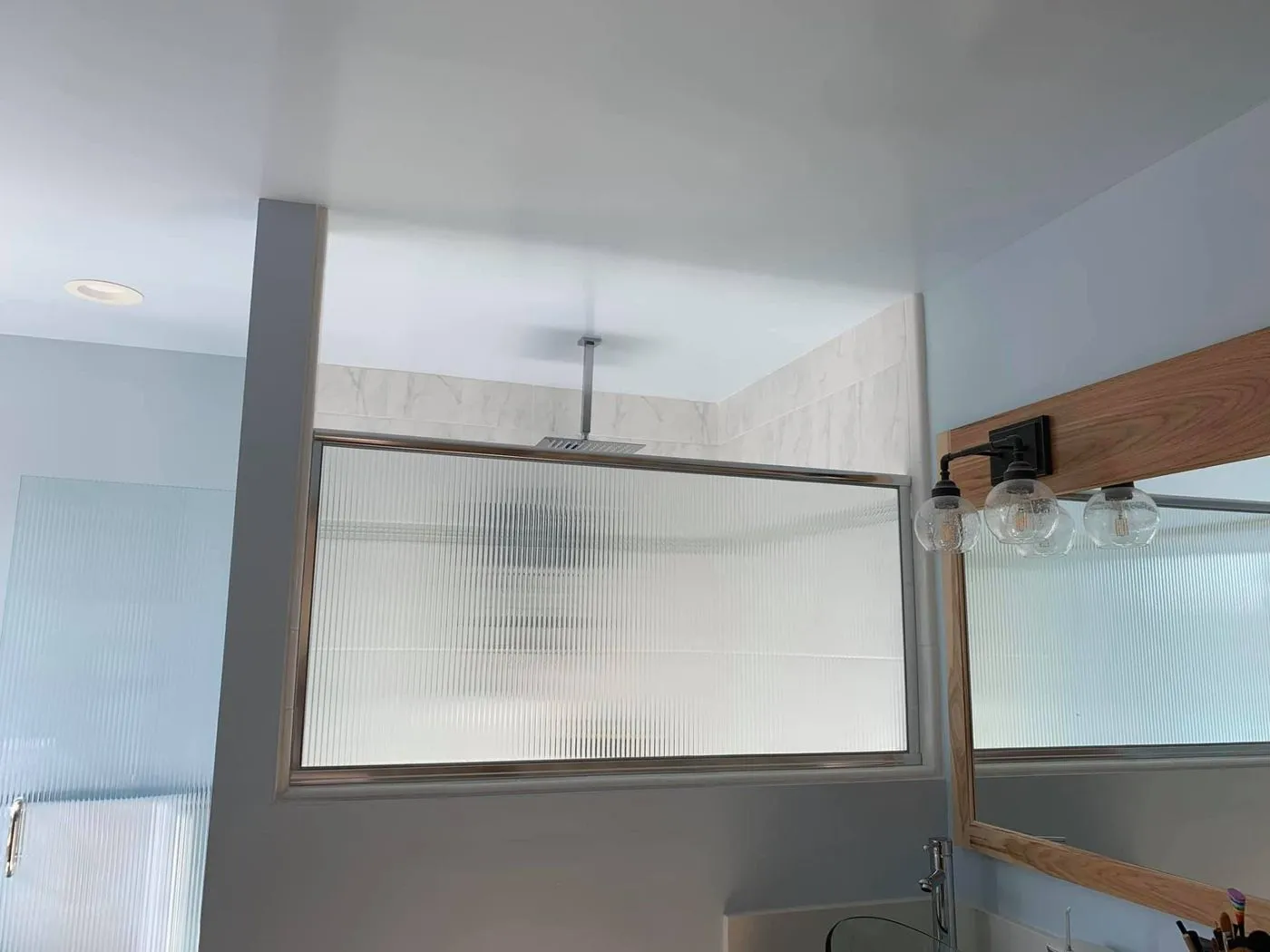 A bathroom with frosted glass panels separating the shower area from the sink and mirror, with a ceiling-mounted showerhead visible above the glass.