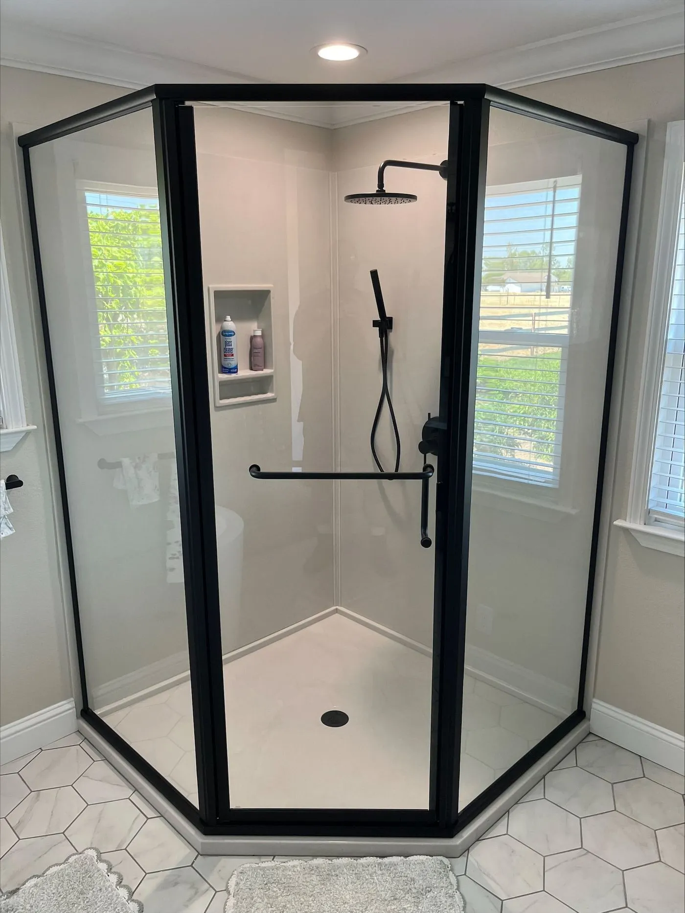Modern hexagonal glass shower enclosure with black frame, overhead rainfall showerhead, wall-mounted hand shower, built-in shelf, and light-colored tile floor.