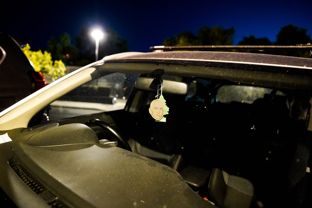 A car interior at night with a cartoon face air freshener hanging from the rearview mirror; the windshield is dusty and a streetlamp is visible outside.