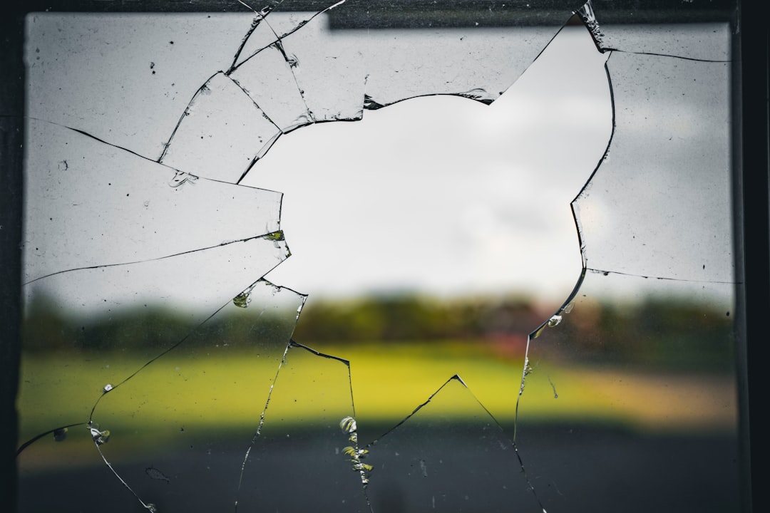 A close-up of a broken window with a large hole and cracks, showing an out-of-focus outdoor scene in the background.