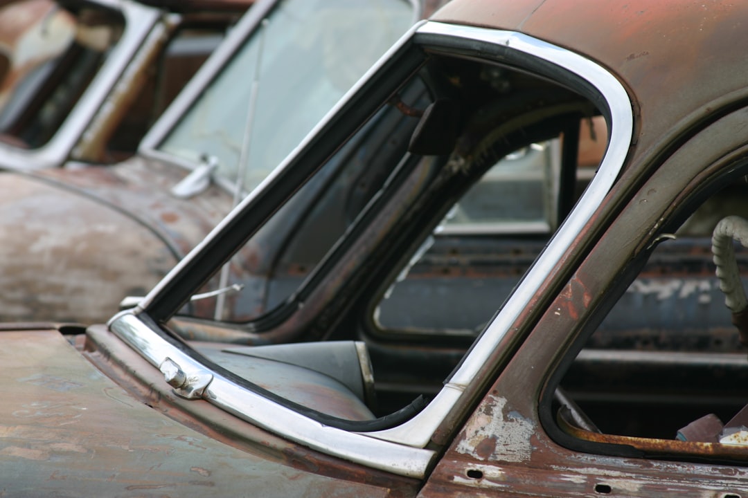 Close-up of the driver side window and roof of a rusty, old car, with more weathered cars in the background.