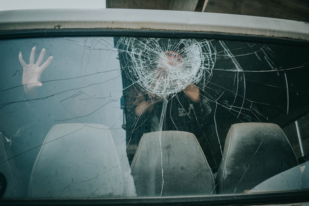 A person’s hand is pressed against a car window with a large, circular crack in the glass; two empty seats are visible inside.