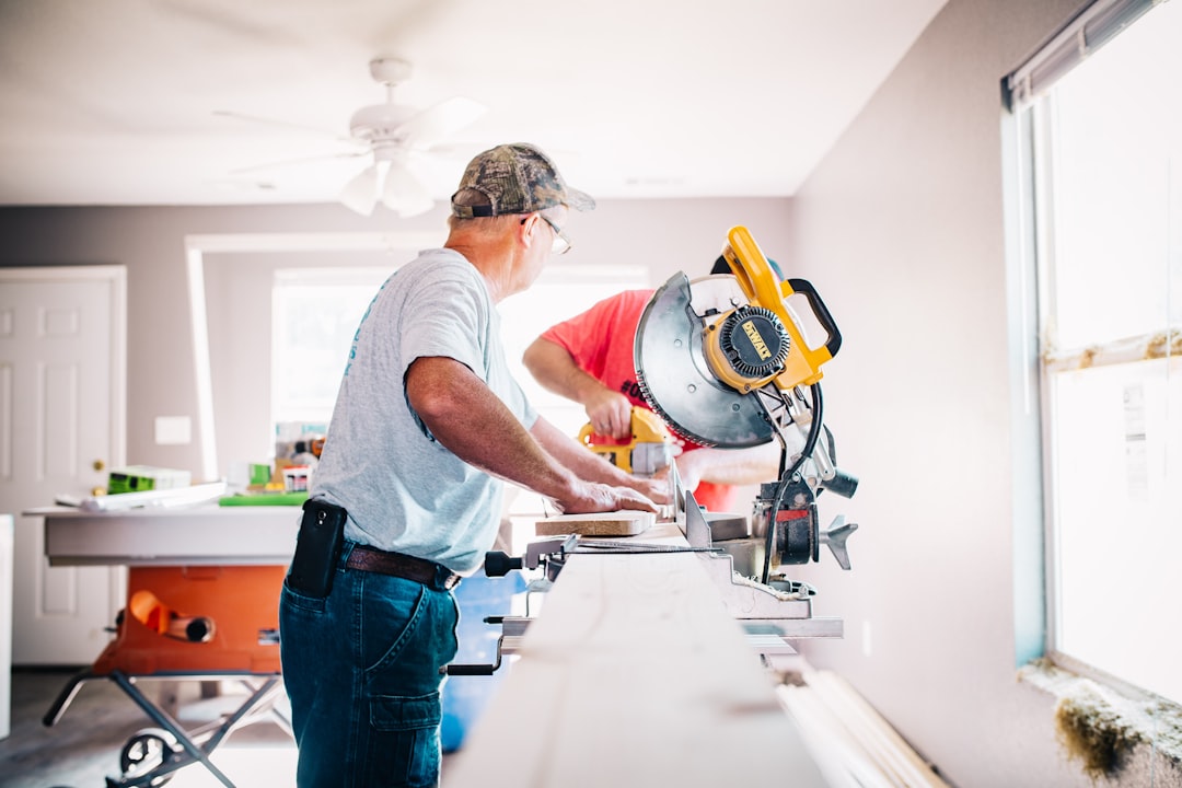 Two men are working indoors with a circular saw, cutting wooden planks on a long workbench near a window. Two men are working indoors with a circular saw, cutting wooden planks on a long workbench near a window.