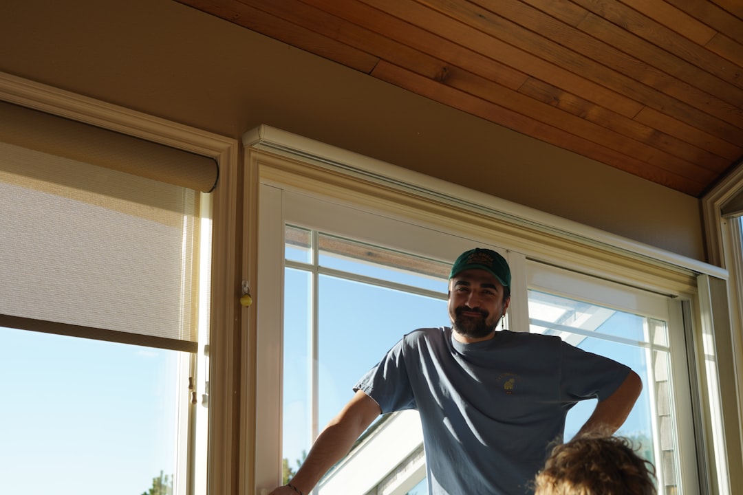 A man in a blue shirt and green cap stands by a large window indoors on a sunny day, with wooden ceiling panels above.