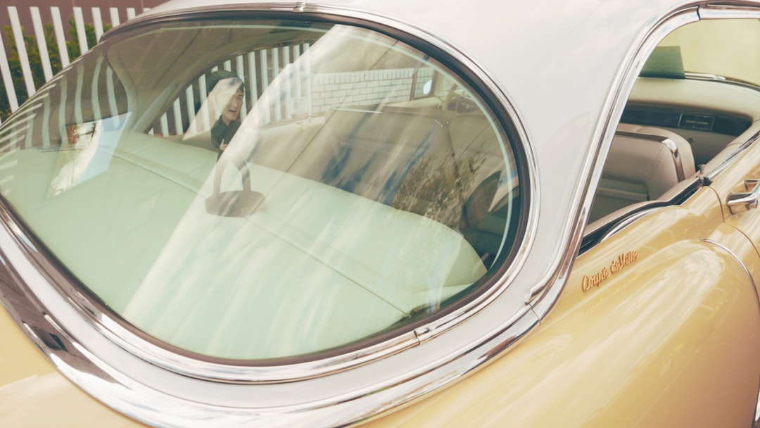 A view through the rear window of a vintage cream-colored car, showing part of the back seat and a reflection of a person outside. A view through the rear window of a vintage cream-colored car, showing part of the back seat and a reflection of a person outside.