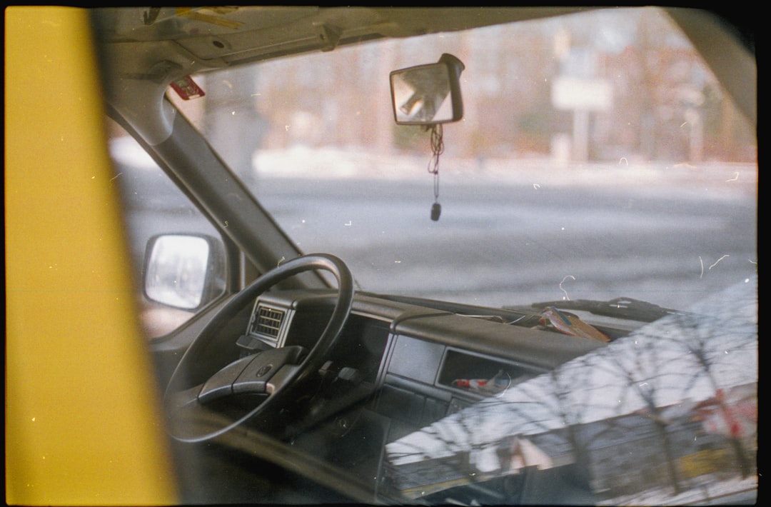 View through a car window showing the steering wheel, dashboard, and a small mirror hanging from the ceiling. Reflections of trees and sky are visible on the glass.