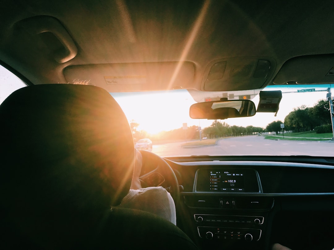 View from inside a car at sunset, showing a driver at the wheel and sunlight shining through the windshield onto the dashboard and road ahead. View from inside a car at sunset, showing a driver at the wheel and sunlight shining through the windshield onto the dashboard and road ahead.