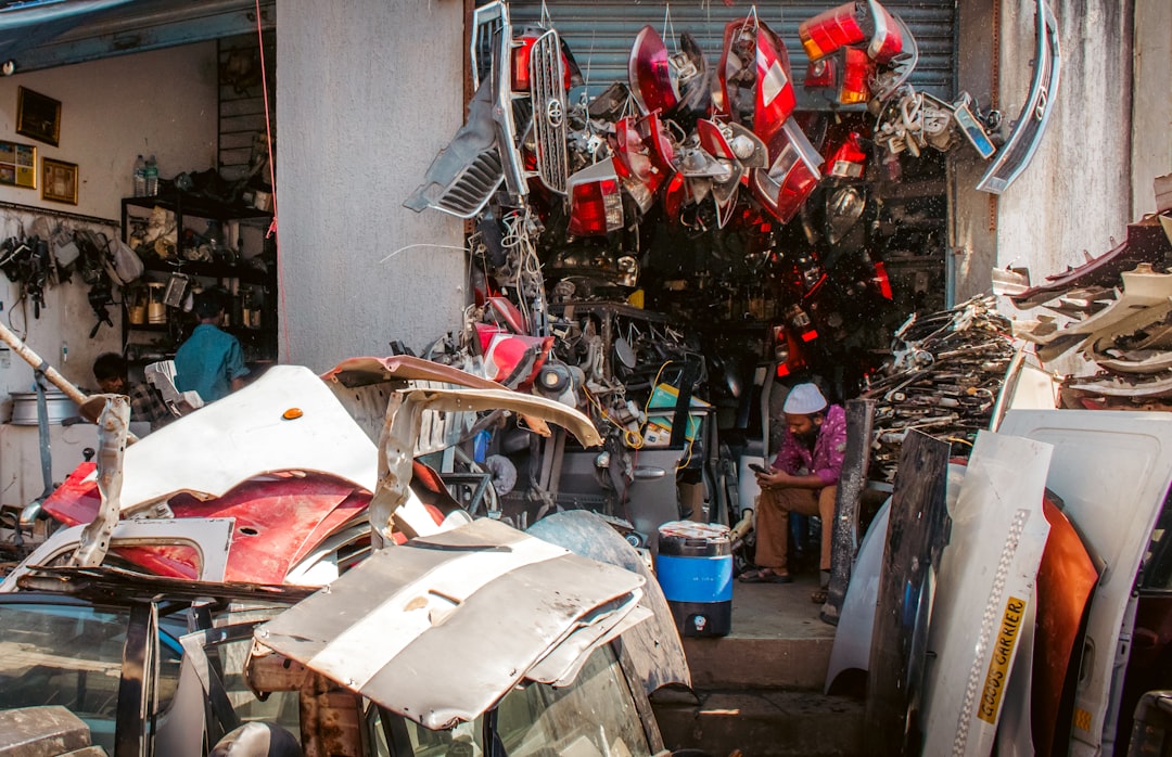 A cluttered auto parts shop with car parts piled outside and hanging above the entrance, and a person sitting inside near the doorway. A cluttered auto parts shop with car parts piled outside and hanging above the entrance, and a person sitting inside near the doorway.