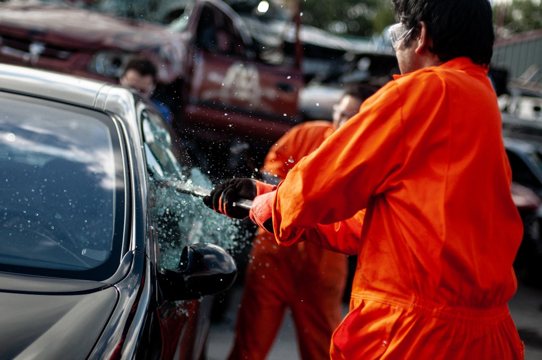 A person in an orange jumpsuit breaks the side window of a car with a tool, while others and stacked vehicles are visible in the background.